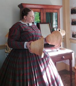 Anna in a red plaid dress looking at two straw bonnets she made during a summer interpretation of women's work in a millinery shop.
